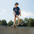 A young boy wearing a helmet and a Tandem Kids Tandem Moon Tee is skateboarding at the edge of a concrete skate park ramp on a sunny day.