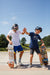 Two teenage boys at a skatepark, high-fiving each other. One boy leans on a graffiti-covered skateboard, while the other holds his skateboard by his side, both wearing helmets and Tandem Kids Tandem Sun Tee.