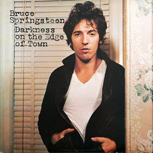 Album cover for "Bruce Springsteen and the E Street Band - Darkness on the Edge of Town" by Bruce Springsteen, featuring the artist leaning against a wallpapered wall with a disheveled look.