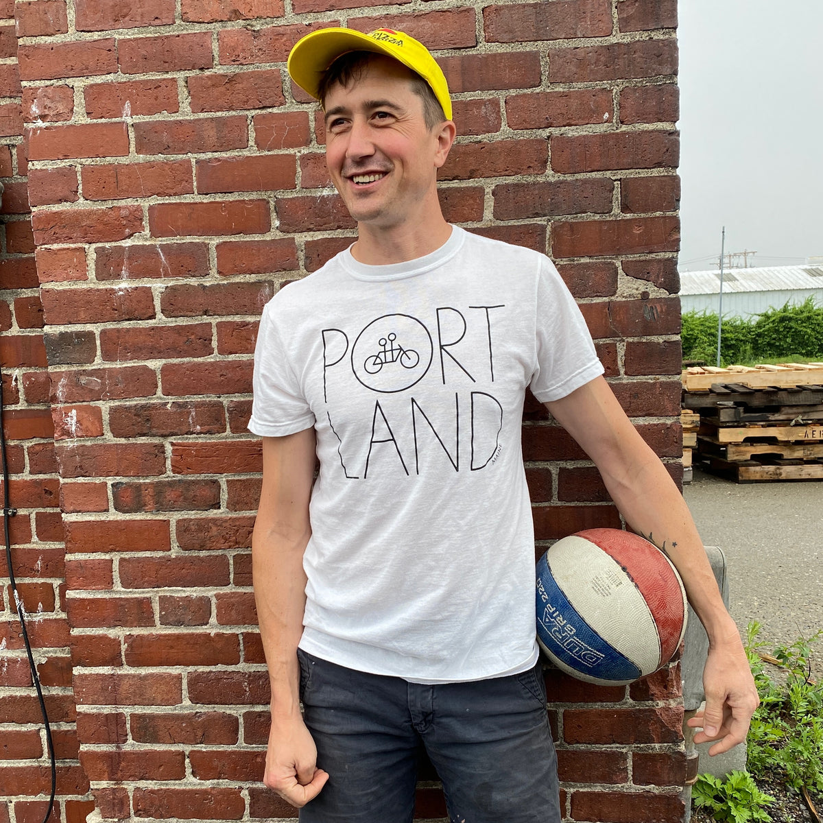 A man in a white Recycled Portland Tee from Tandem Coffee Roasters smiles by a brick wall, holding a basketball under his arm and wearing a yellow cap.