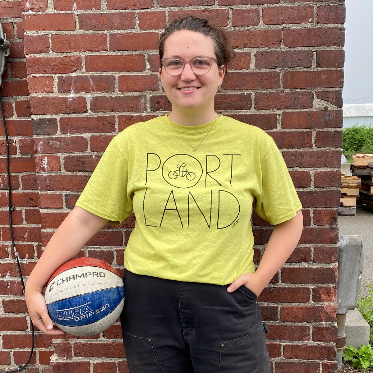 A smiling woman wearing glasses and a bright yellow Recycled Portland Tee made from upcycled cotton with Tandem Coffee Roasters printed on it, stands in front of a brick wall holding a football.