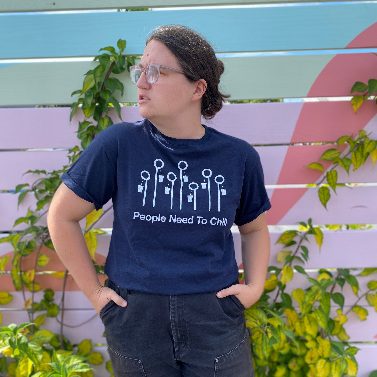 A young woman in glasses, smiling and giving a thumbs up, wearing a navy blue Tandem Coffee Roasters "People Need To Chill" Tee in front of a colorful striped background with greenery.