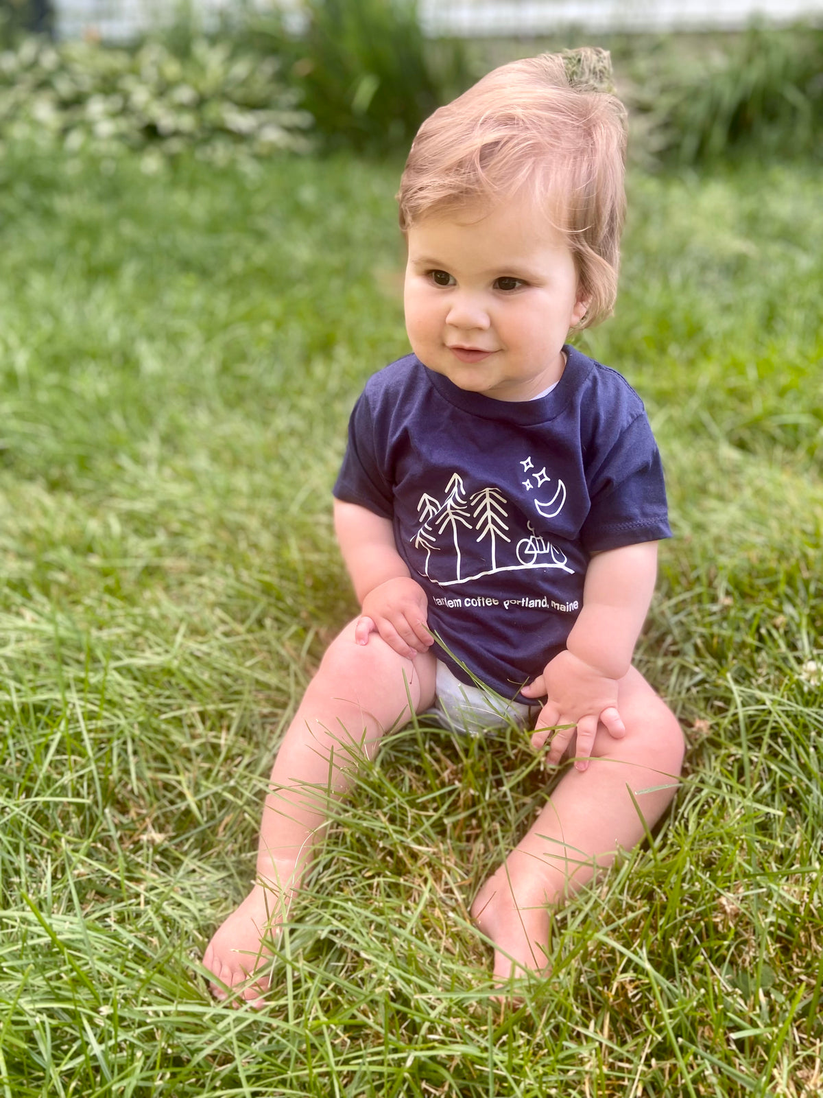A toddler with light hair wearing a Tandem Kids Tandem Moon Tee sits on grass, looking off to the side. The shirt has a graphic of mountains and trees with text. Soft focus background with greenery.