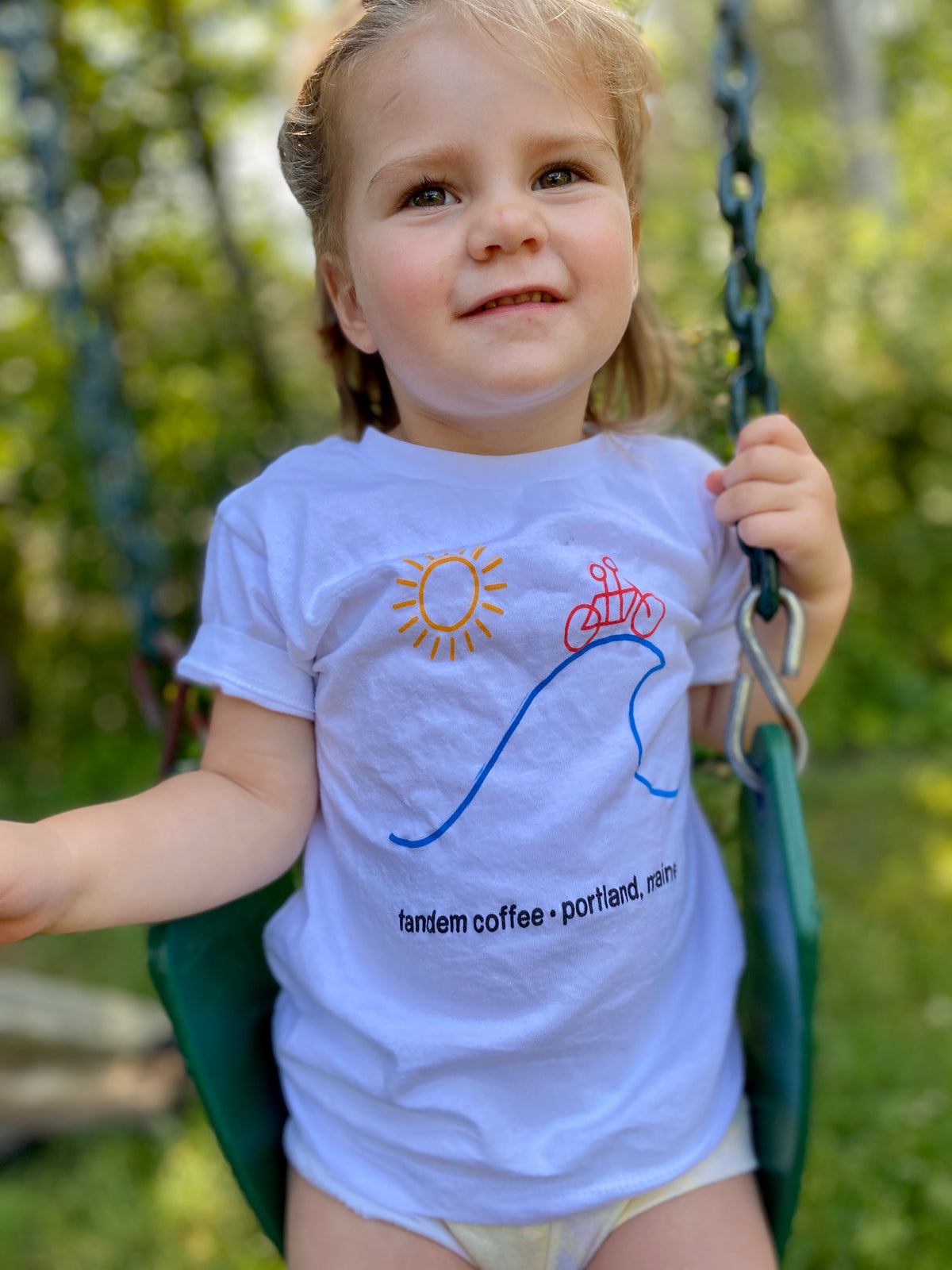 A young child with light brown hair wearing a 100% cotton Tandem Kids Tandem Sun Tee and smiling gently while sitting on a green swing in a sunlit garden.