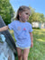 A young girl stands outdoors near a wooden structure, wearing a white 100% cotton Tandem Kids Tee with a Kids Tandem Sun Tee design. She gazes thoughtfully to the side, with her