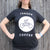 A woman in a black Tandem Coffee Roasters Circle Logo Tee featuring the words "tandem coffee" and a graphic of a tandem bicycle stands against a wooden backdrop.