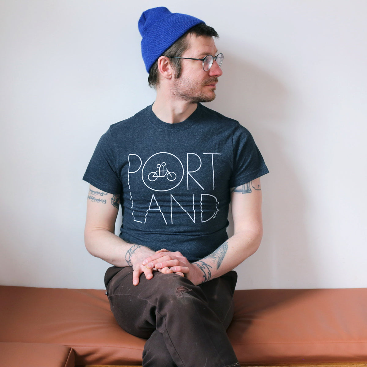 A man wearing glasses, a blue beanie, and a dark t-shirt made of recycled cotton with "Portland" written on it sits on a brown bench, looking to the side with a Tandem Coffee Roasters Recycled Portland Tee.