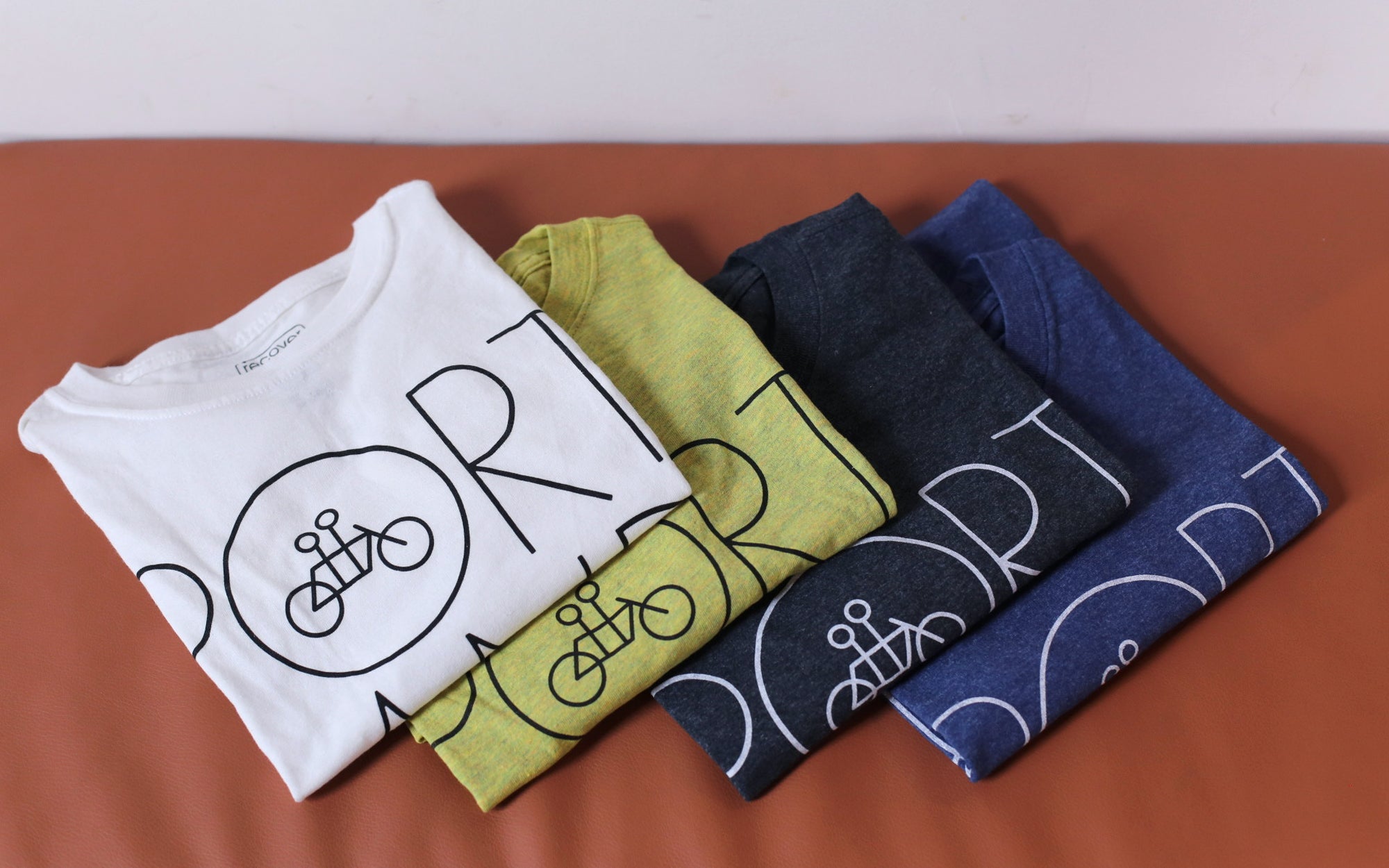 Four neatly folded Recycled Portland Tee t-shirts in white, yellow, green, and blue made of upcycled cotton with the word "sport" and icons of a person cycling printed on an orange surface by Tandem Coffee Roasters.