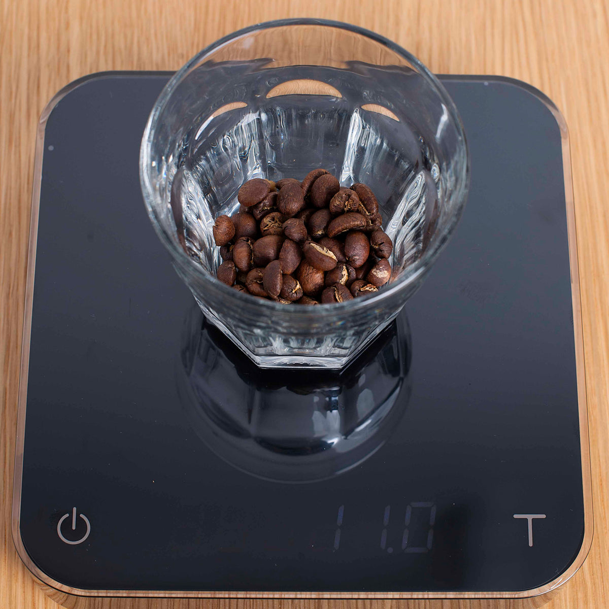 A clear glass cup filled with coffee beans, resting on a USB rechargeable Tandem Acaia Pearl Black scale showing a weight of 111 grams, placed on a wooden surface.